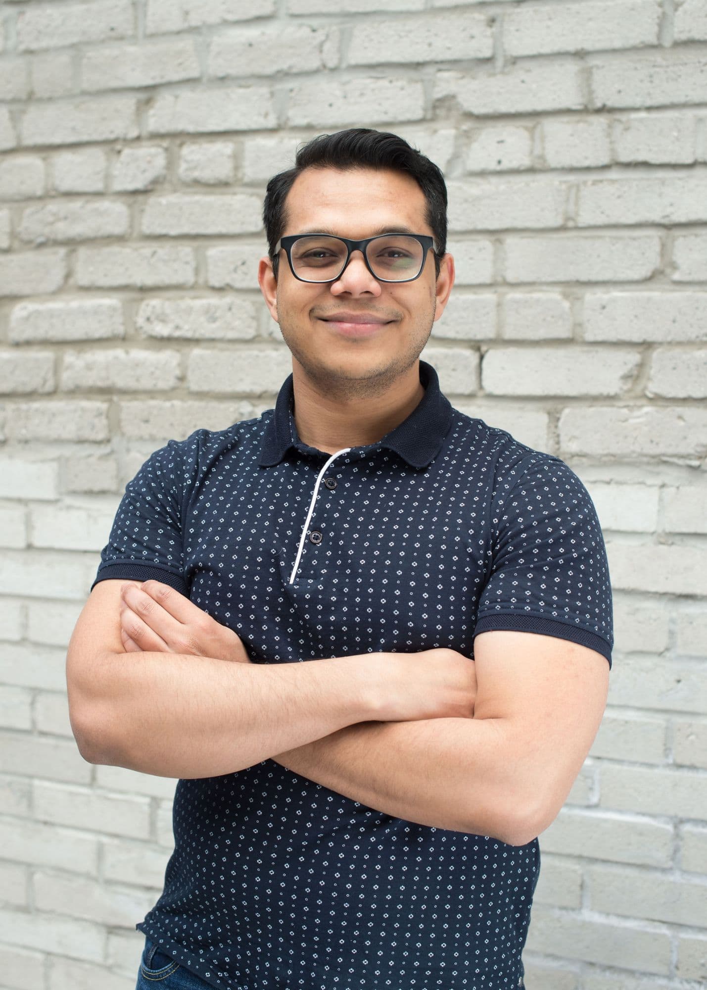 Portrait of Kowsheek, arms crossed, smiling, in front of a pale brick wall